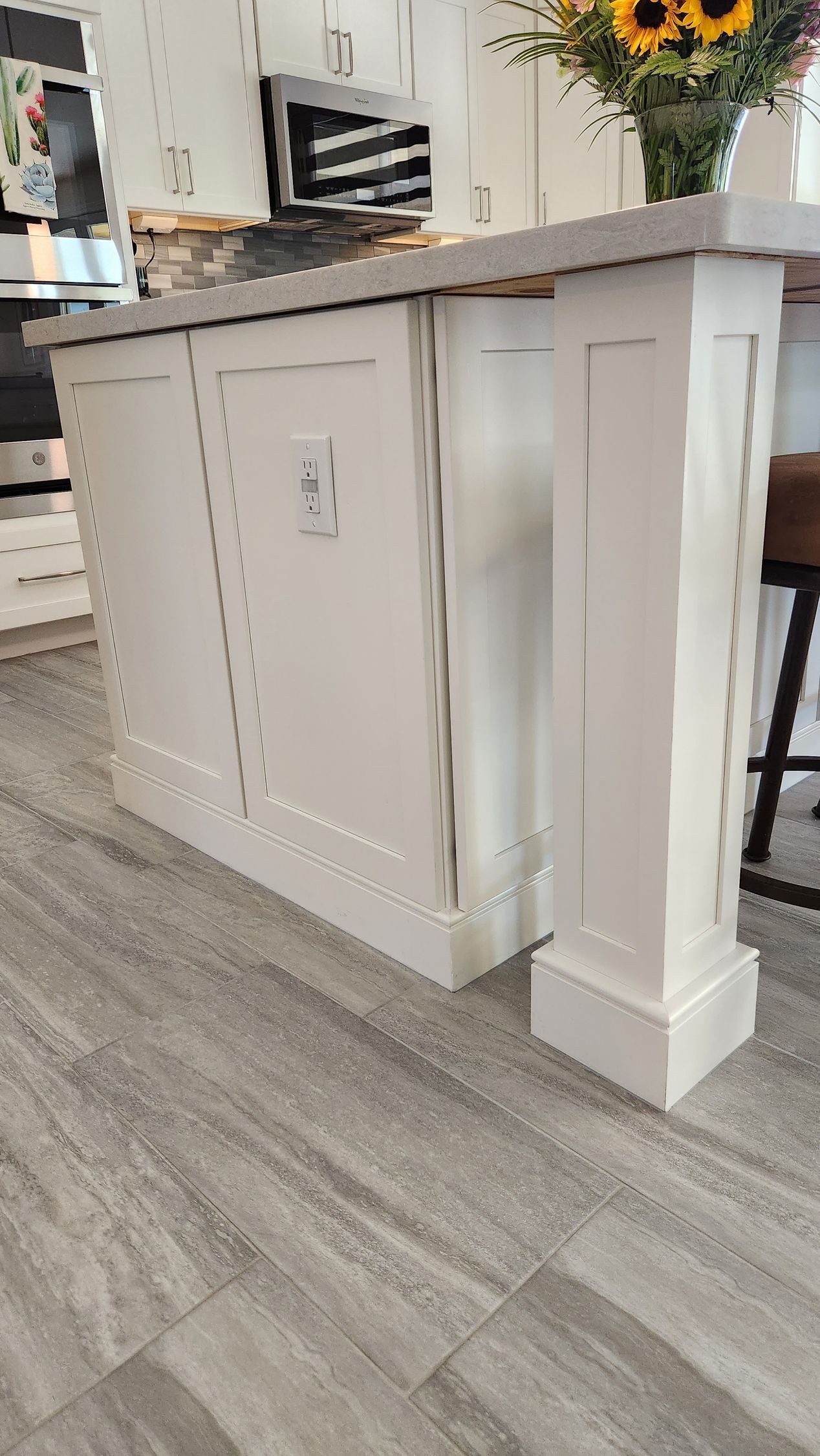 White kitchen island with decorative paneling and a countertop, shown on a gray wood floor with sunflowers on top
