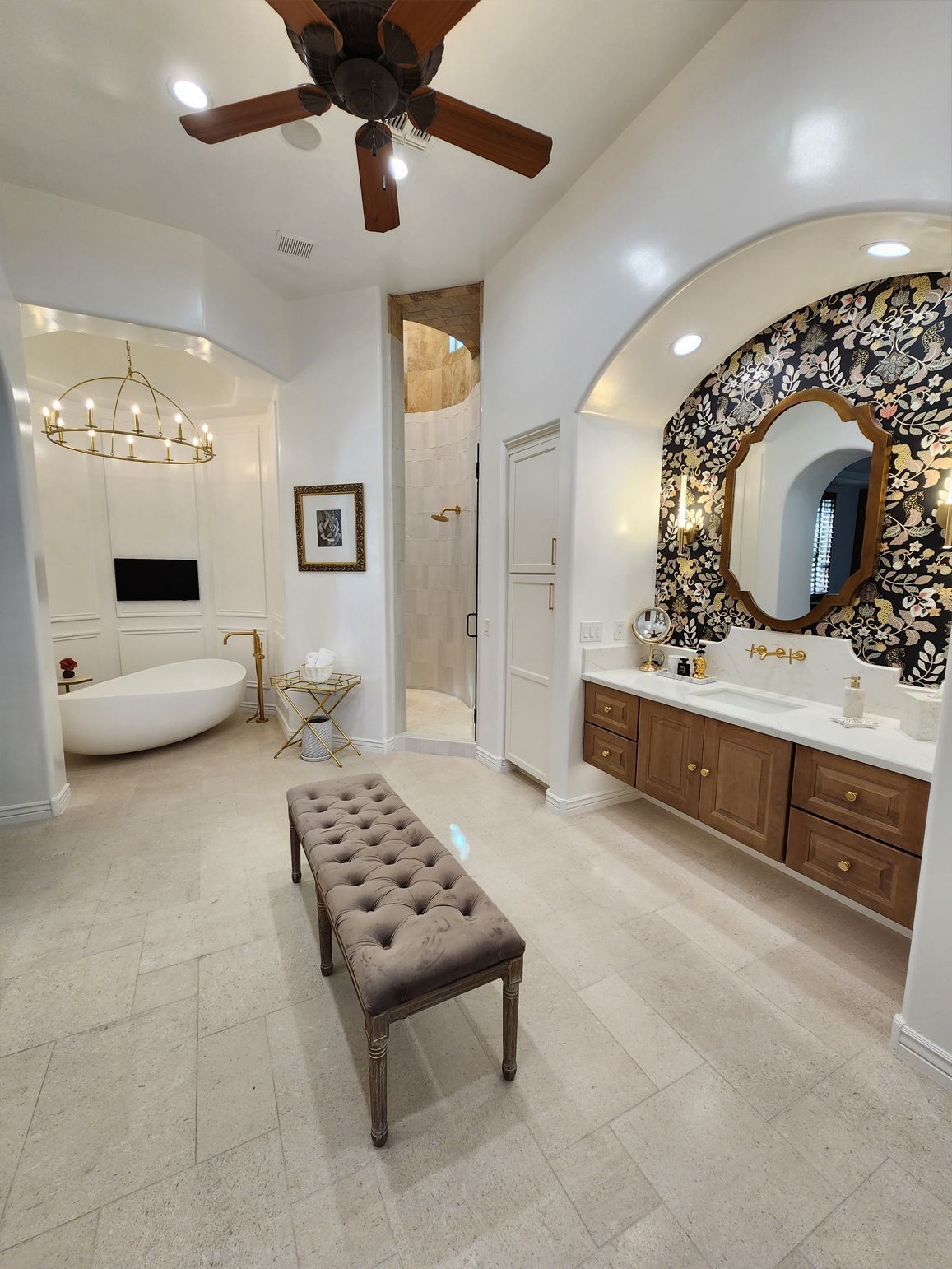 Bright bathroom with soaking tub, vanity, bench, large ornate mirror, and ceiling fan under arched ceiling.