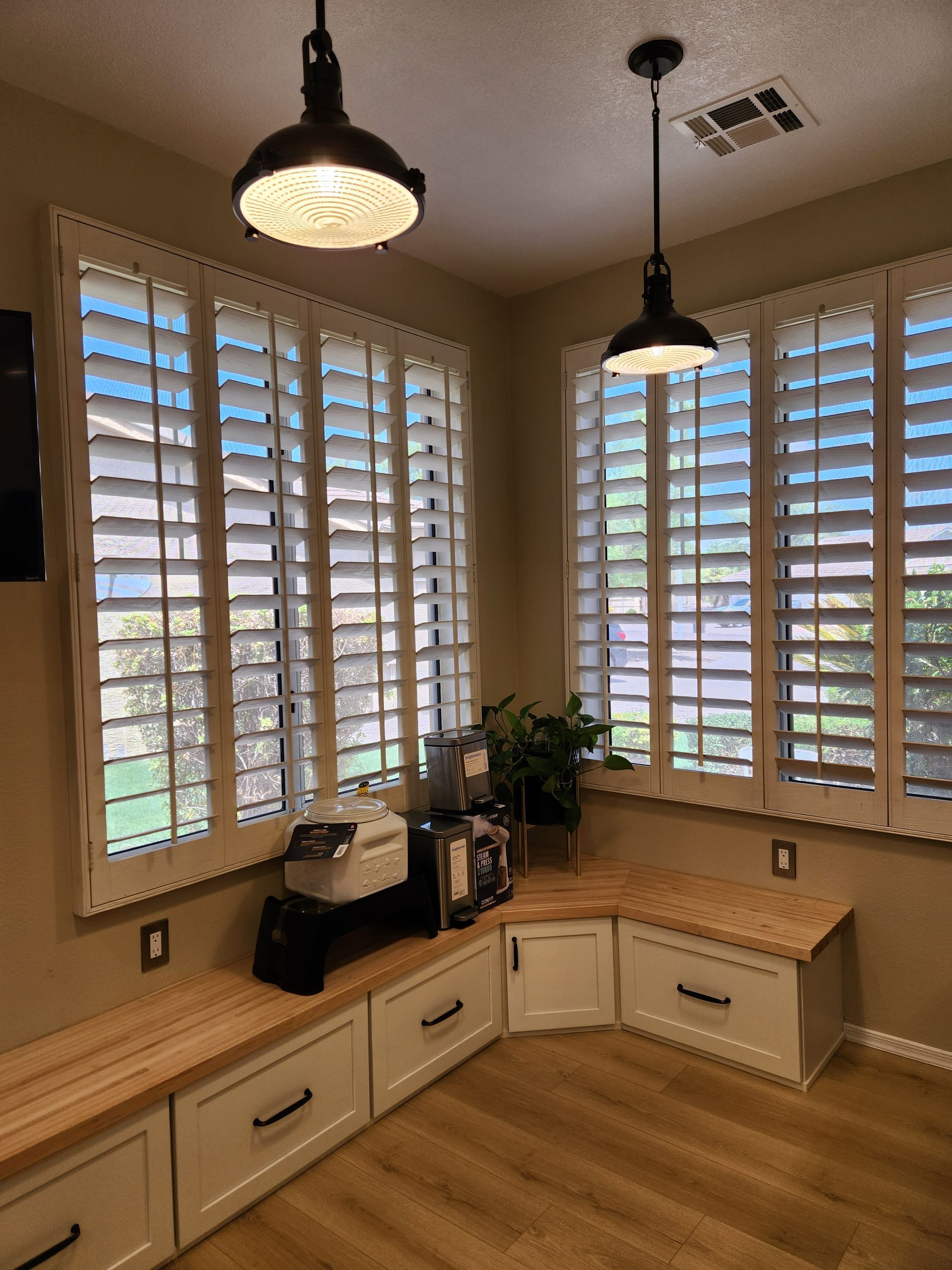 Bright corner office nook with white shutters, built-in desk, hanging lights, and wood flooring.