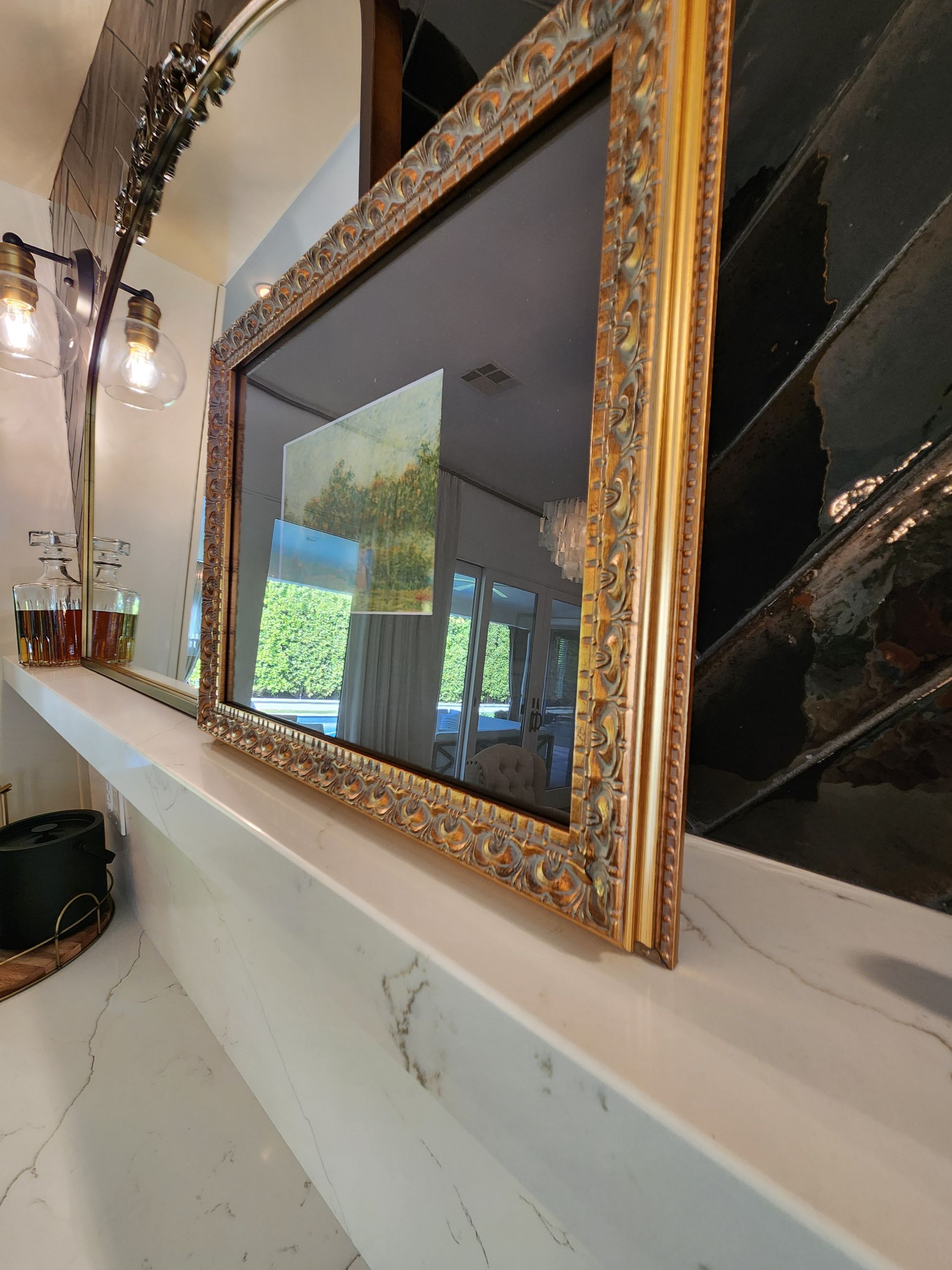 Gold-framed mirror on a white mantel, reflecting a bright window and greenery in a modern room