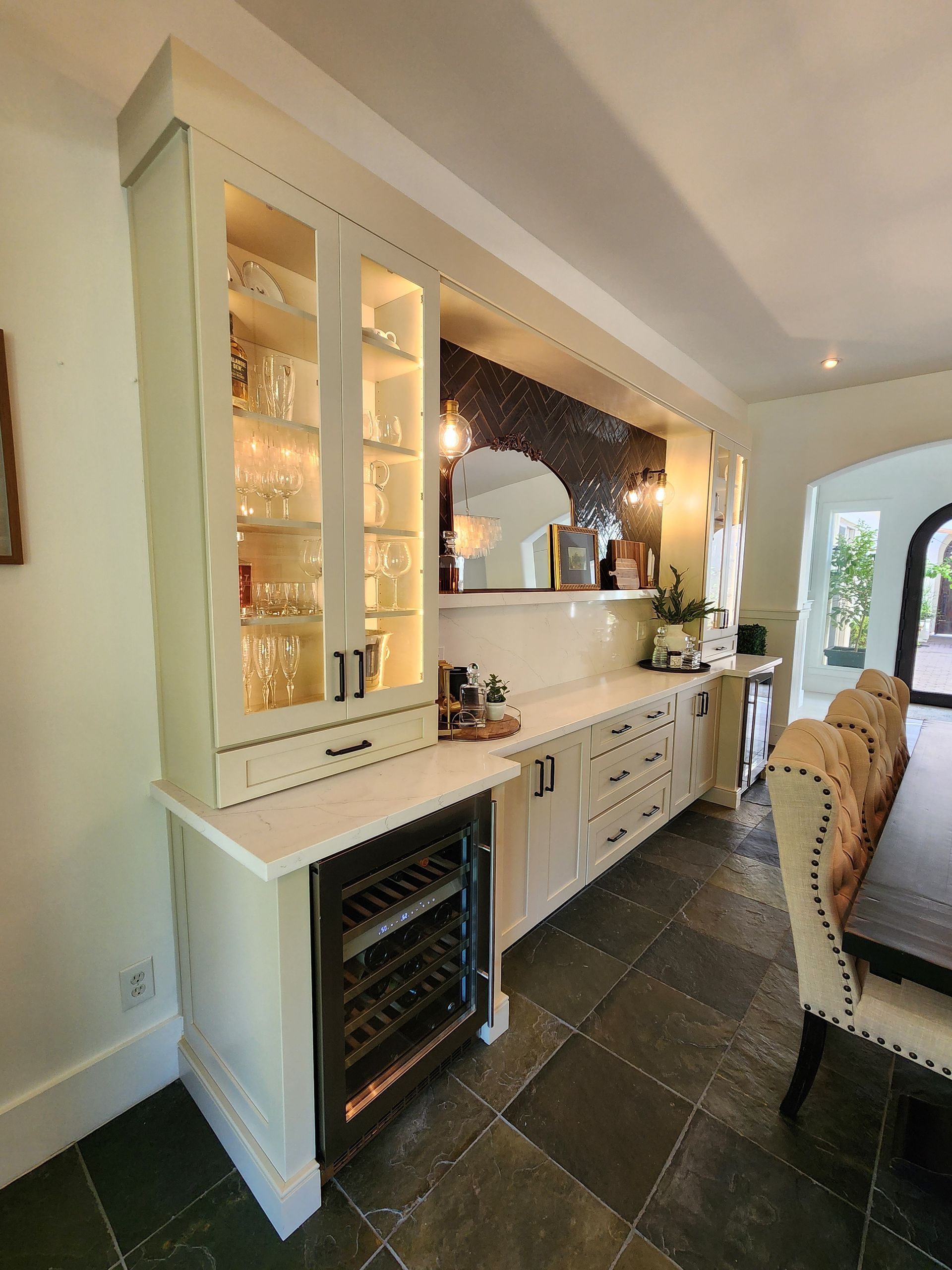 Bright kitchen bar area with glass cabinets, wine fridge, and white cabinetry beside a dining table