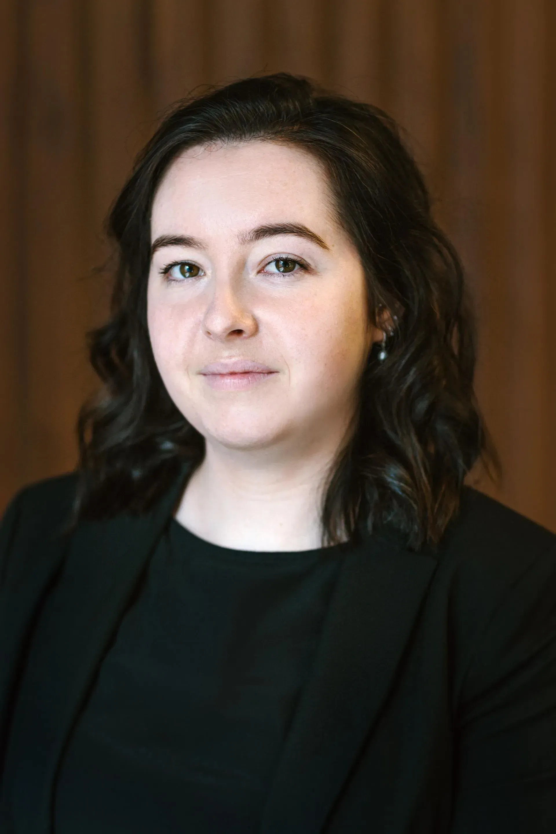 Woman with dark hair smiles in black blazer. Brown backdrop.
