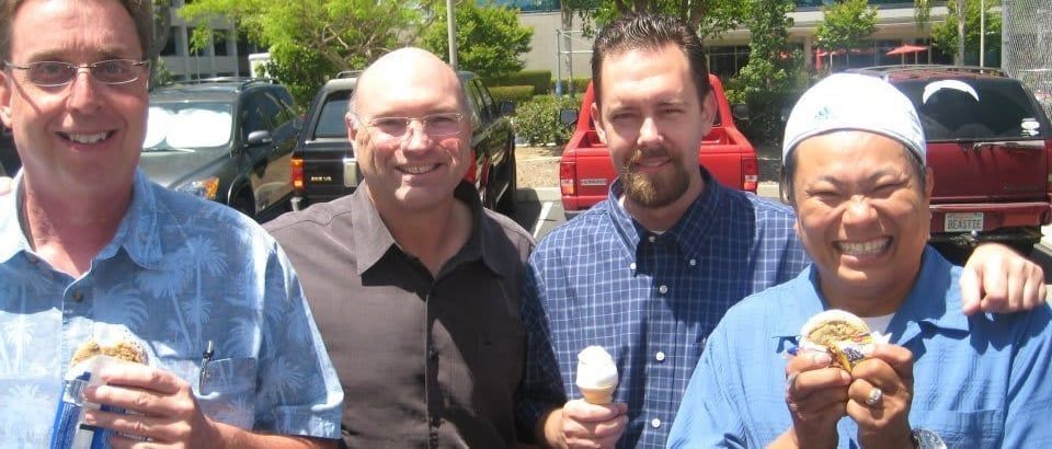 Four people smiling outdoors, each holding ice cream cones in front of parked cars.