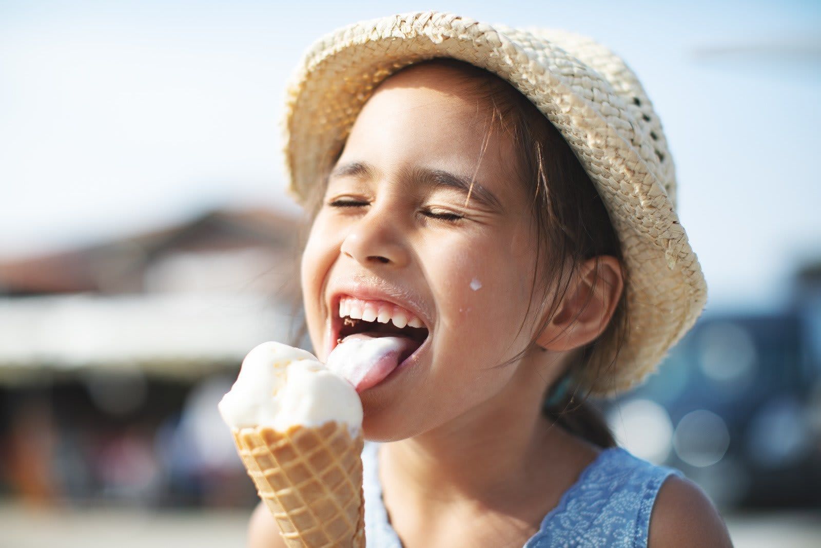 Child licking an ice cream cone outdoors, wearing a straw hat and smiling in bright sunlight