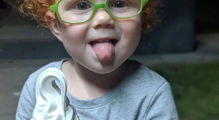 Child with green glasses sticking out their tongue while holding a white shoe.