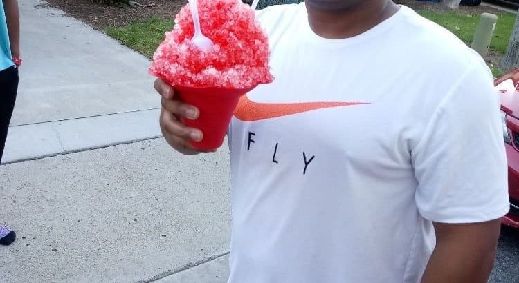 Person in a white Nike shirt holding a red shaved ice cup outdoors on a sidewalk.