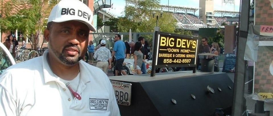 Man in white BIG DEV cap stands beside a large black smoker at an outdoor barbecue stall