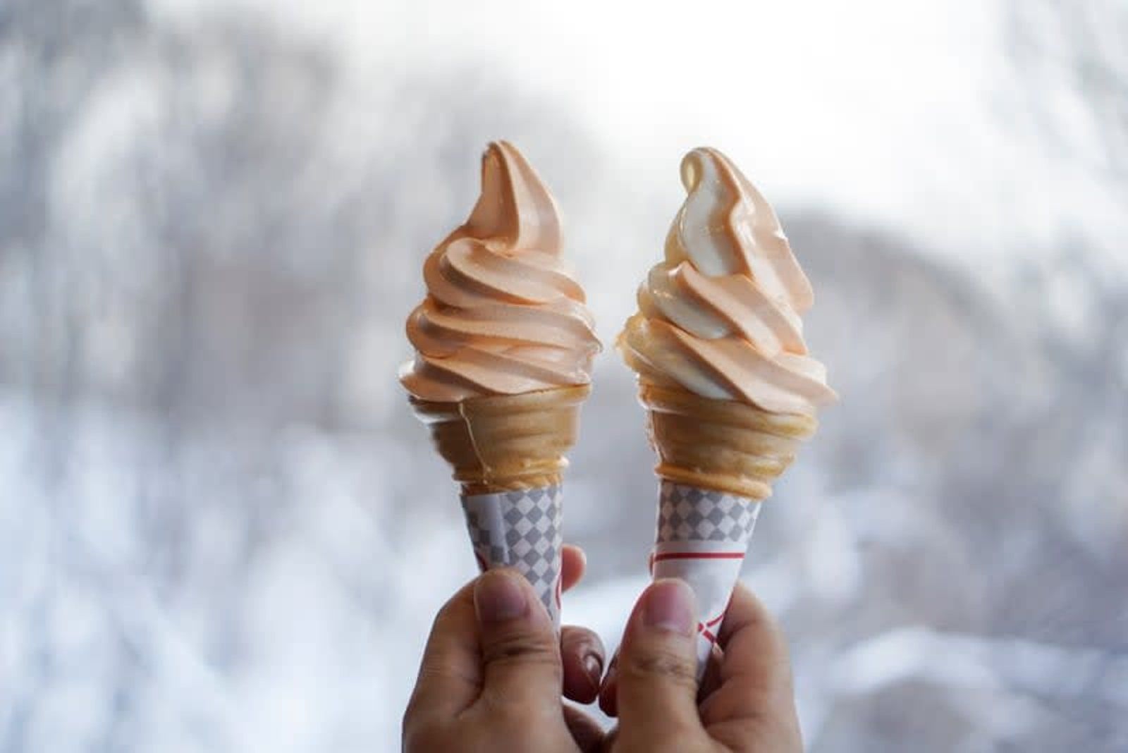 Two soft-serve ice cream cones held up against a snowy outdoor background