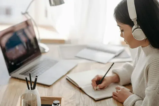 Person wearing headphones writes in a notebook beside a laptop at a bright desk.