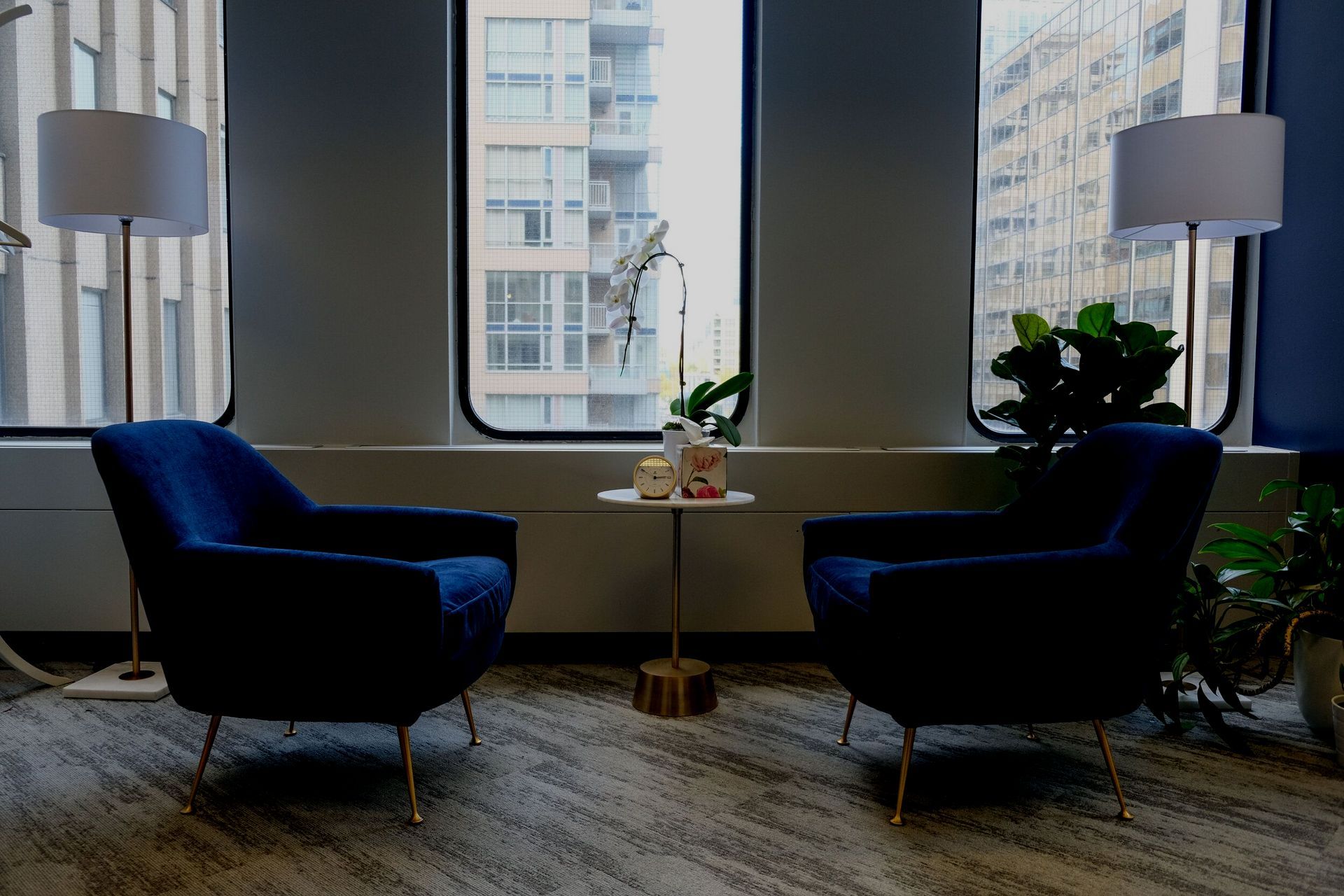 Modern lounge with two blue chairs, a small table, floor lamps, and city windows.