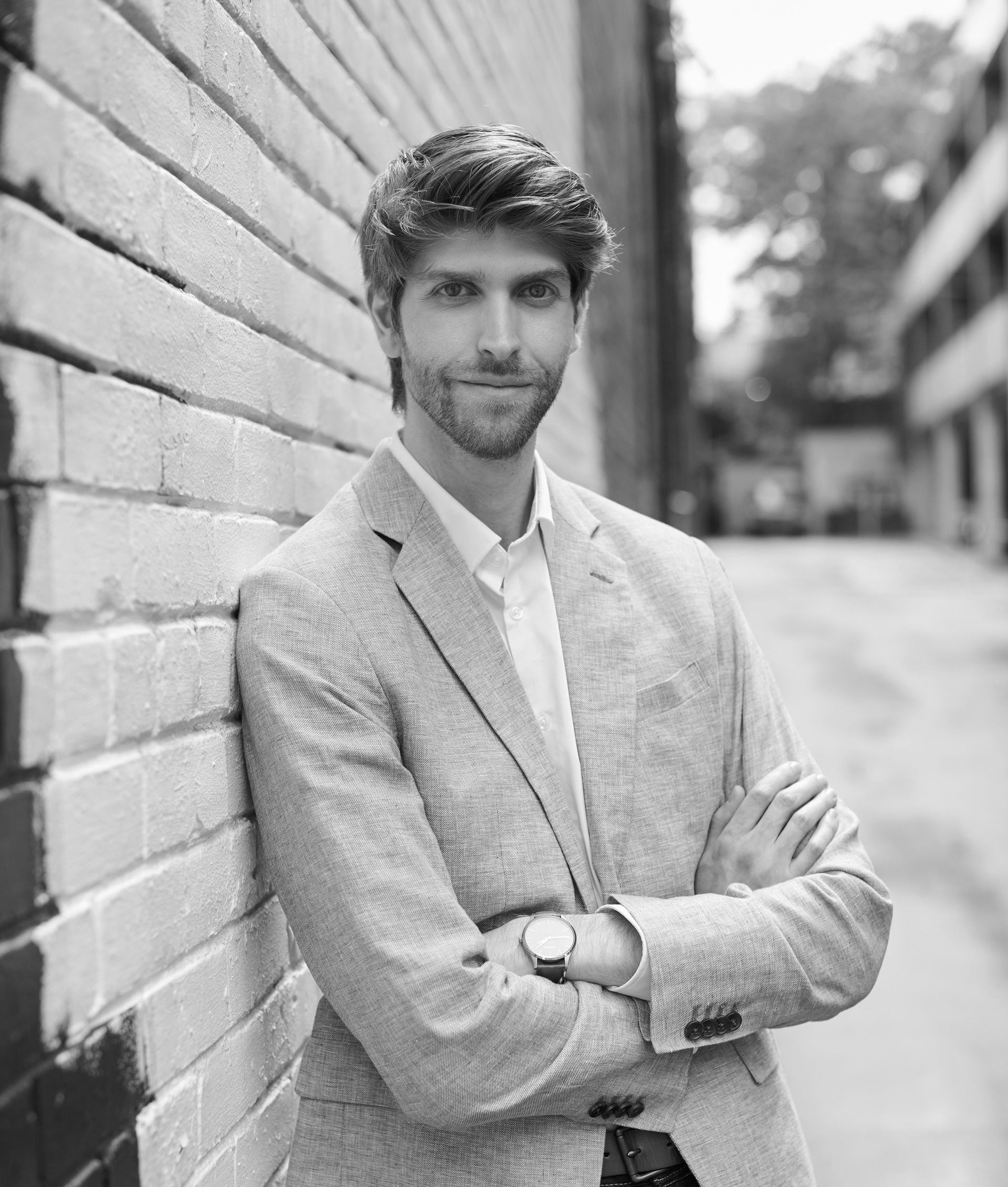 Man leaning against a brick wall on a city street, arms crossed, wearing a light blazer.