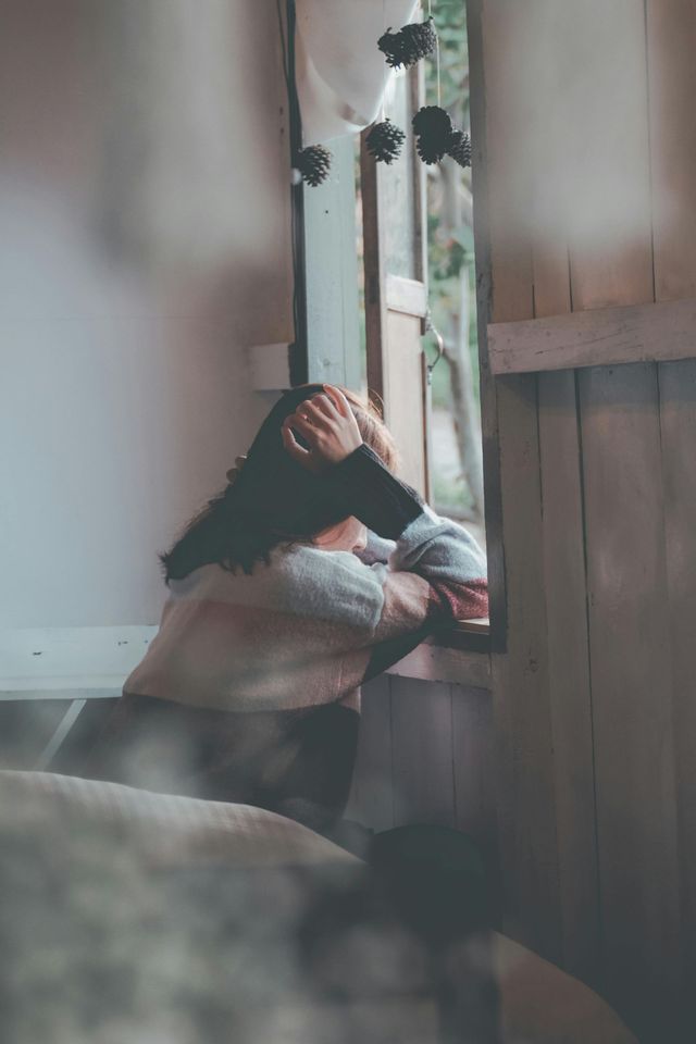 Person sitting by a window, resting their head on a pillow in soft indoor light.