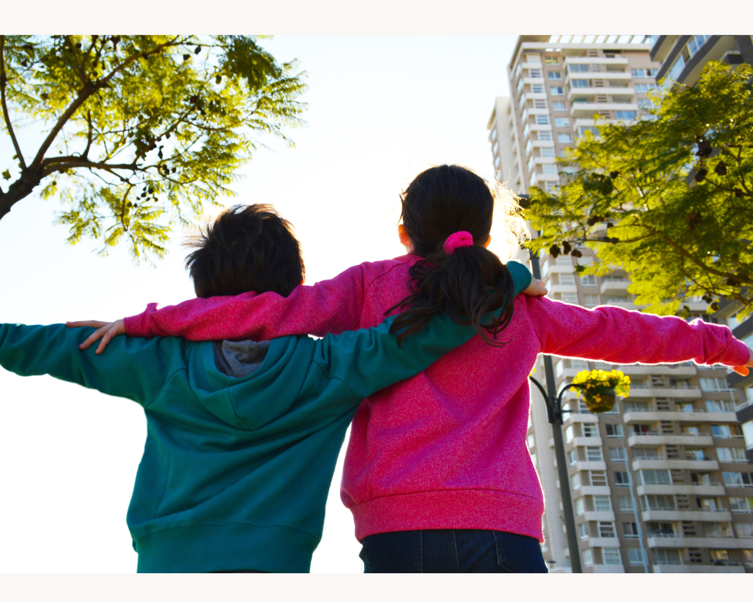Two children with arms outstretched in a park, with trees and tall buildings in the background.