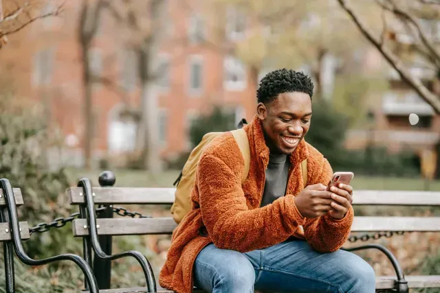 Person in an orange jacket smiling on a park bench while looking at a phone