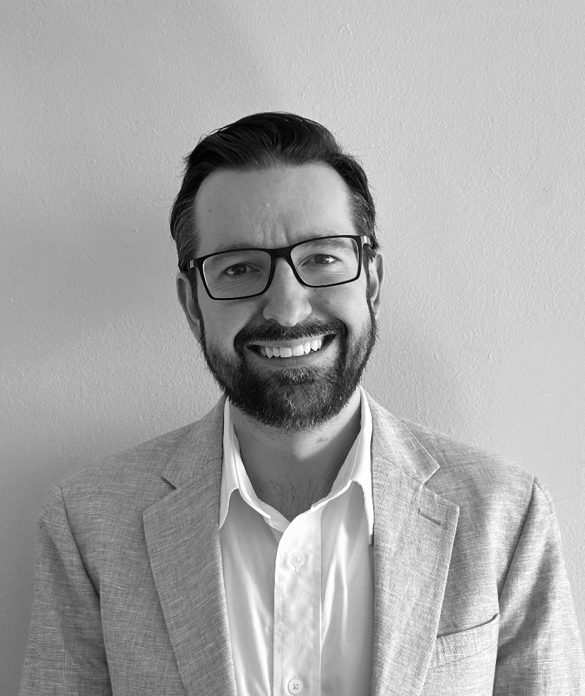 Smiling man with glasses in a light blazer and white shirt against a plain wall