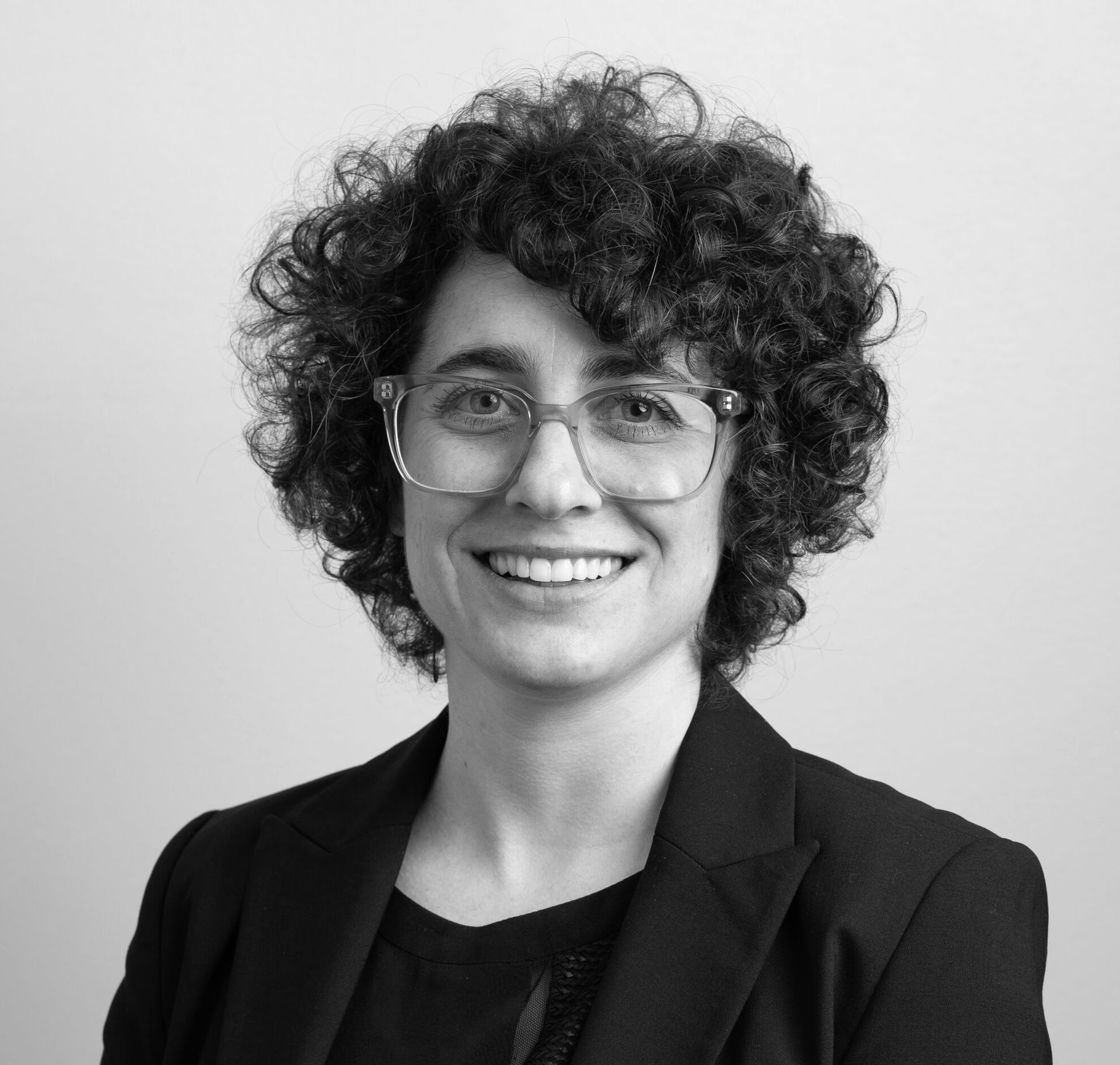 Portrait of a smiling person with curly hair and glasses in a dark blazer