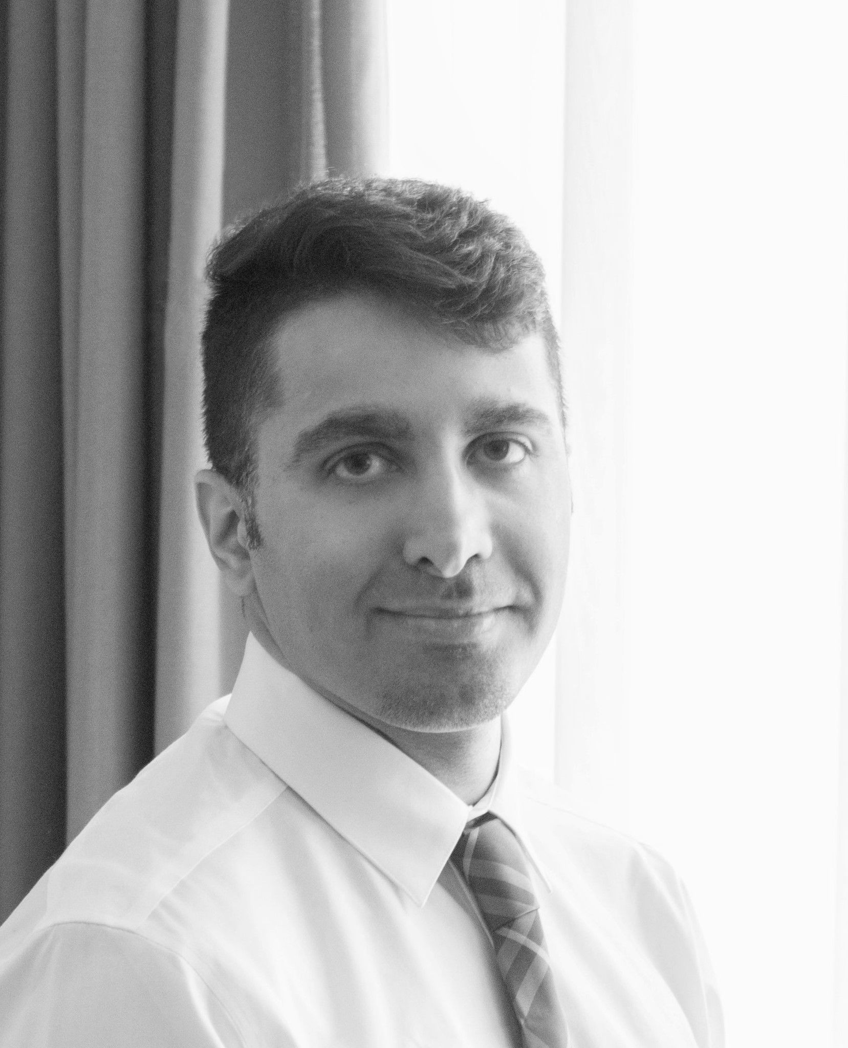 Person in a white shirt and tie, standing indoors by a curtain, looking at the camera.