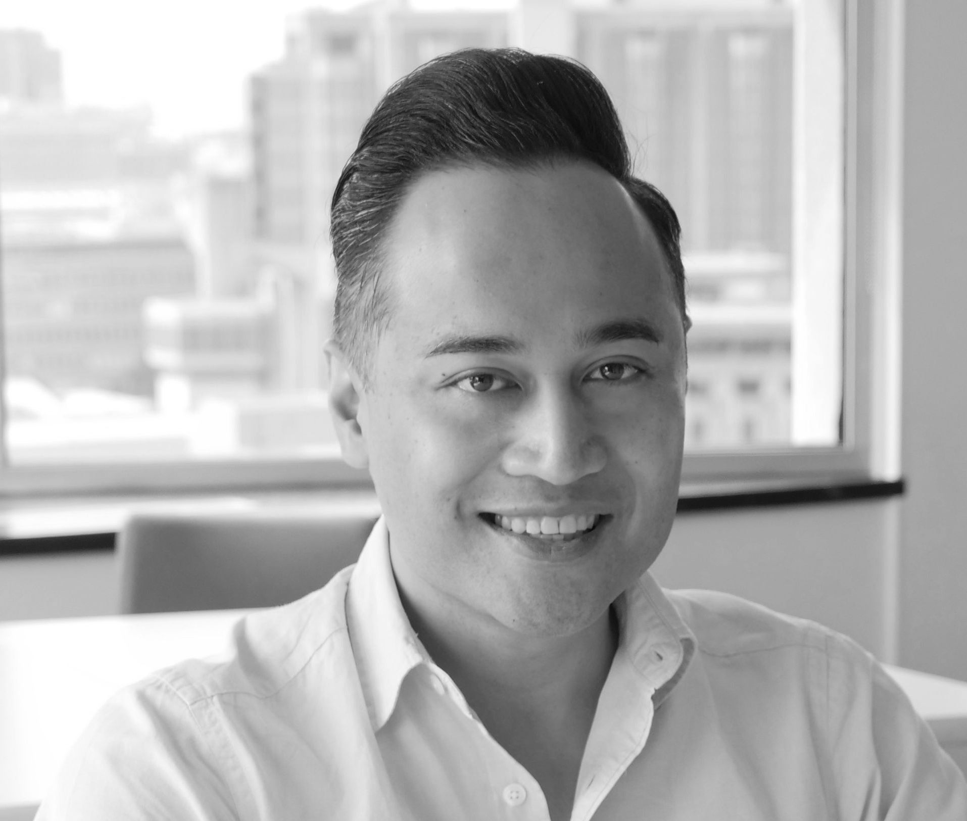 Smiling person in a white shirt seated indoors with a city view in the background