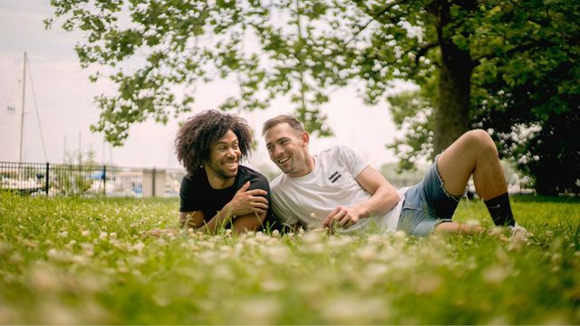 Two people lying on grass and smiling under a tree in a sunny park