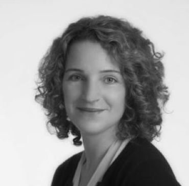 Black-and-white studio portrait of a smiling person with curly hair and a dark blazer.