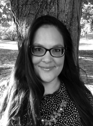 Woman with glasses smiling outdoors among trees in black and white.
