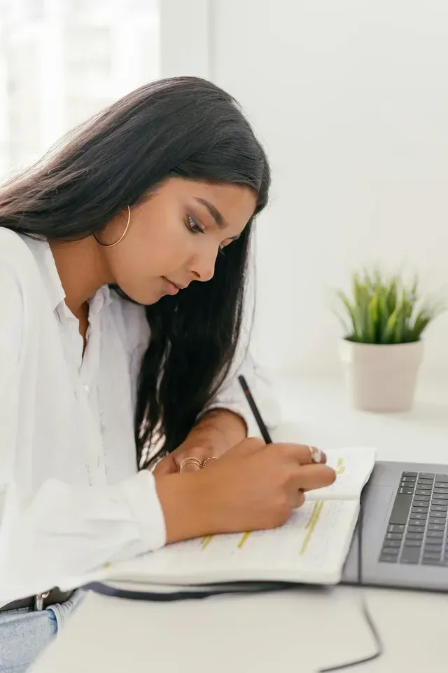 Woman writing in a notebook beside a laptop on a bright desk