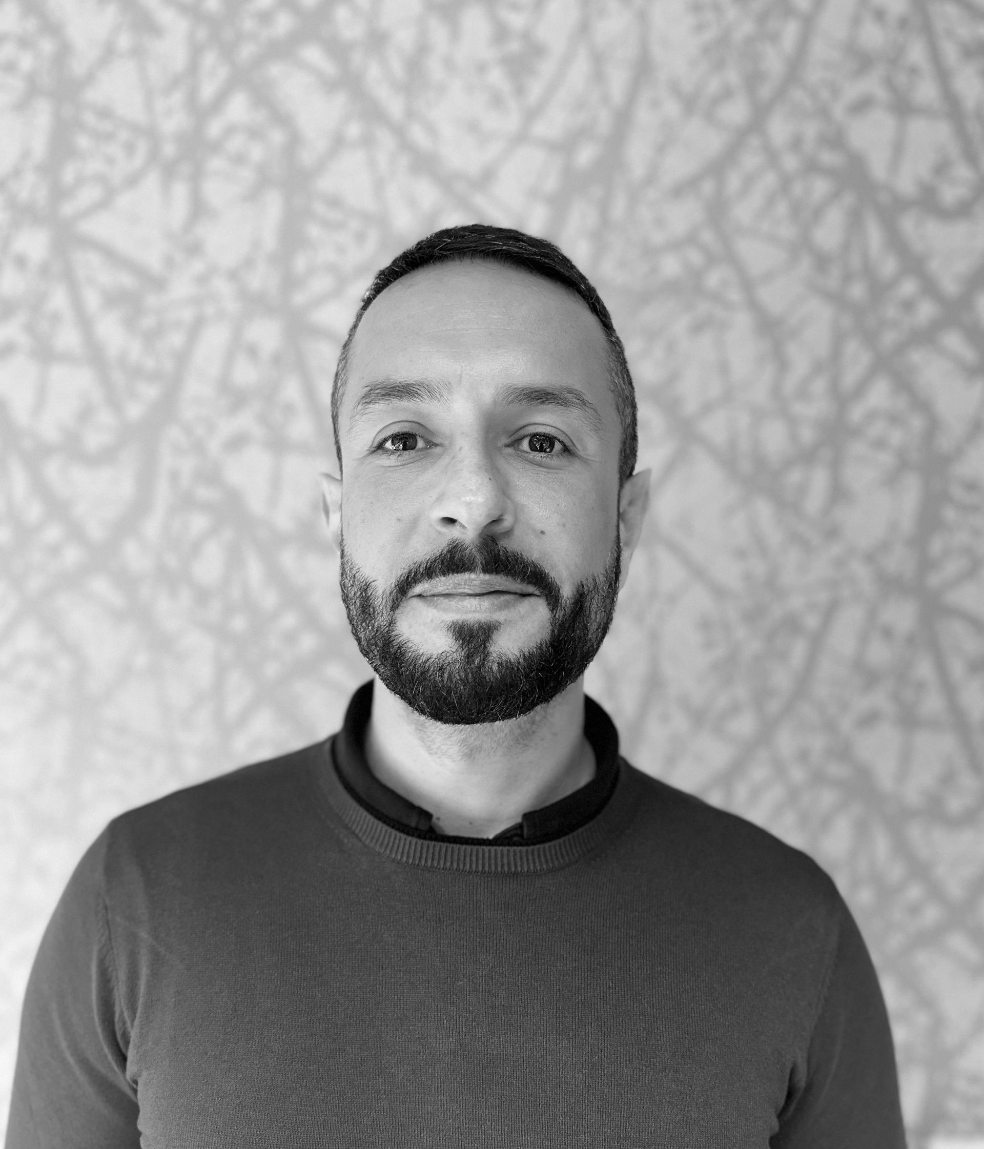Portrait of a man in a dark sweater, standing against a light patterned wall.
