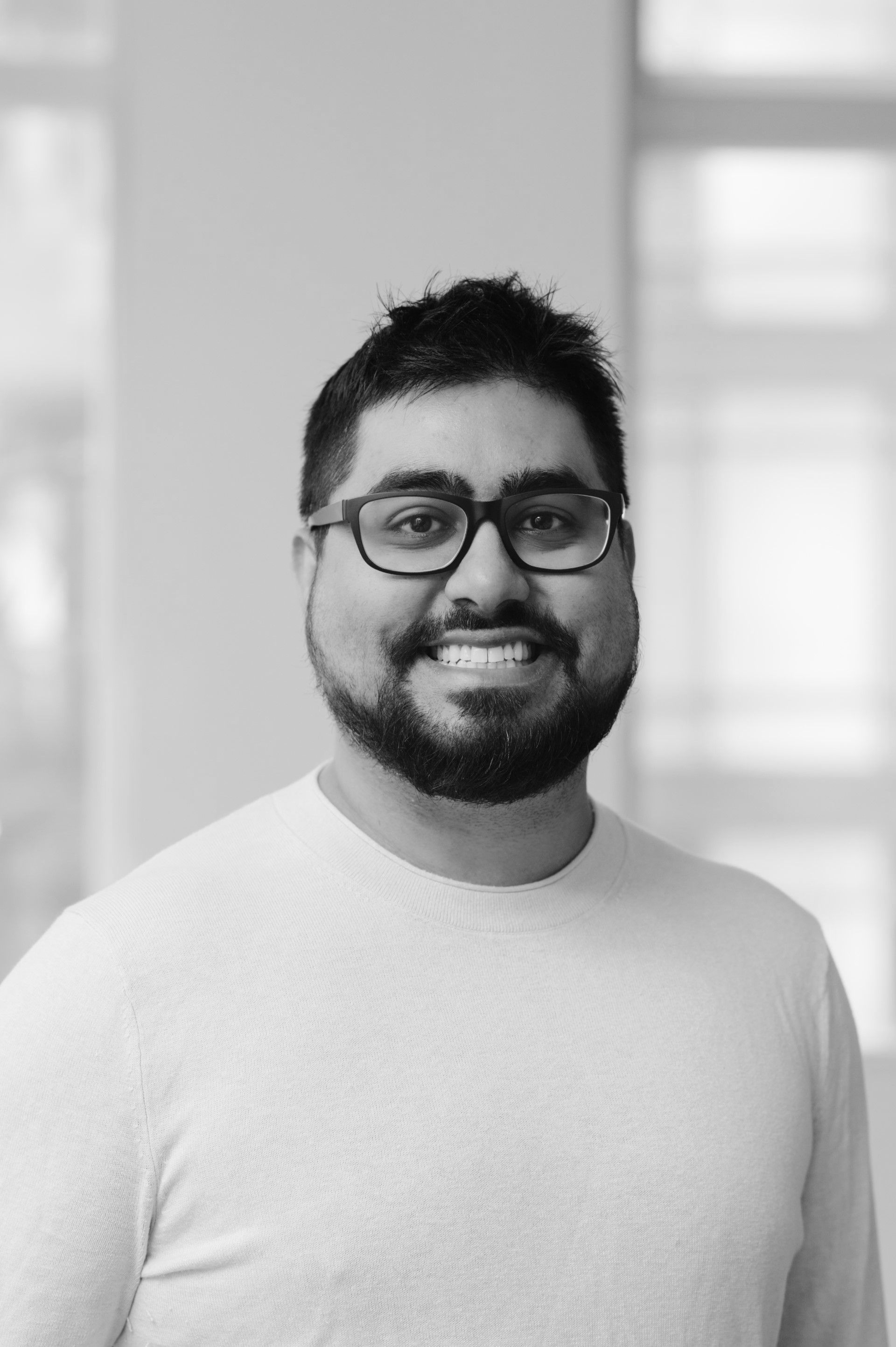 Smiling person with glasses and beard, wearing a light sweater against a bright indoor background