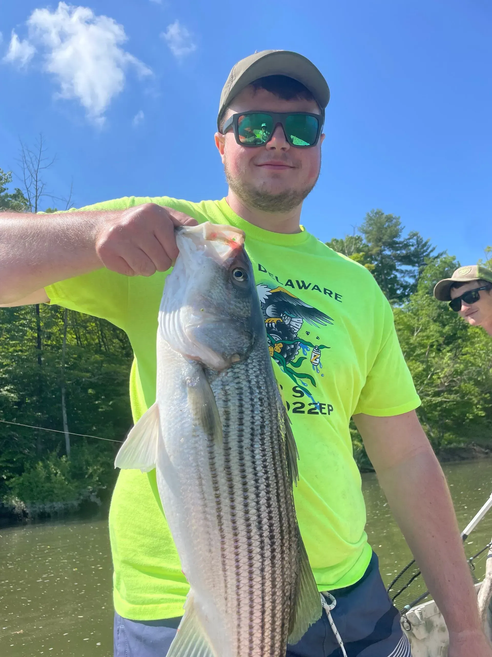 Ready Scout Aquatic Engineer, Andrew Hess, in a bright yellow shirt and sunglasses holds a large striped bass while on a boat outdoors.