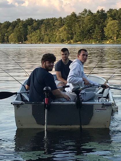 Three people fishing from a small boat on a calm lake at sunset, with trees along the far shore.