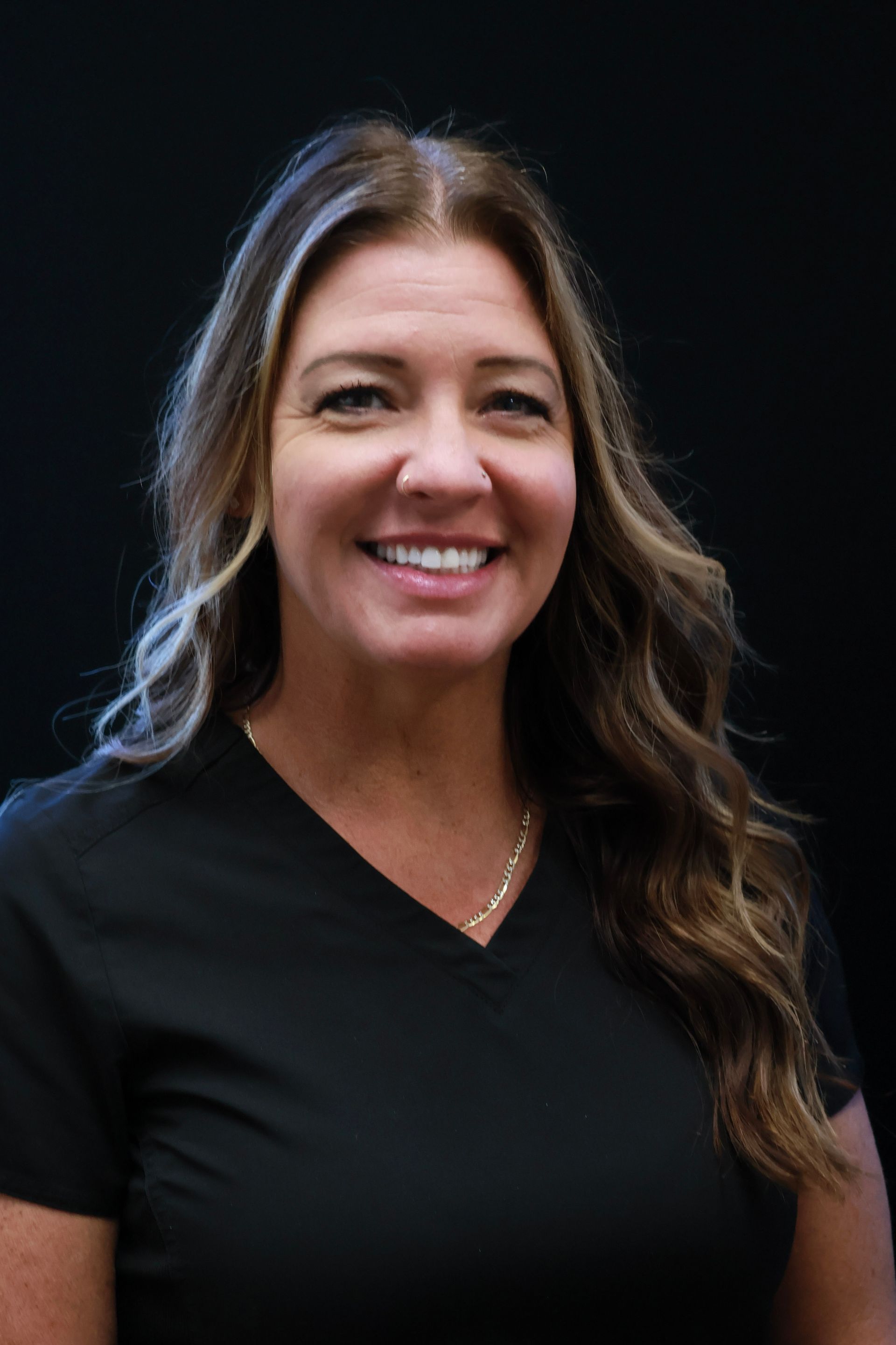 Woman in black v-neck shirt smiles at the camera, wavy brown hair, dark background.