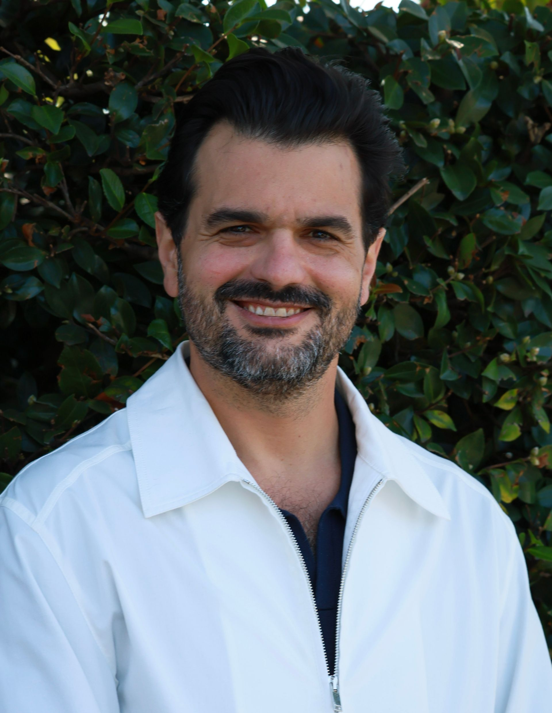 Man with dark hair and beard, smiling, wearing white jacket, standing outdoors in front of green foliage.