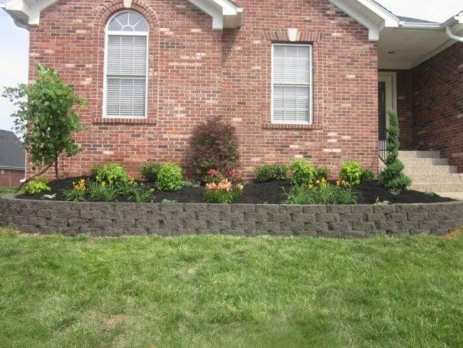 Brick house with a raised garden bed filled with plants and flowers, bordered by a dark retaining wall, set against a green lawn.