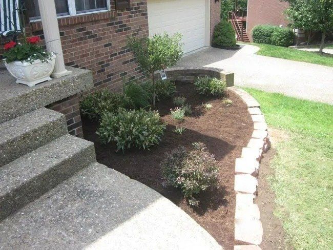 Brick home with a landscaped garden bed; plants sit in front of the house by the driveway and steps.