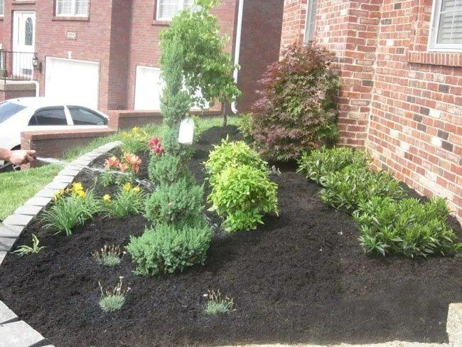 A landscaped garden bed in front of a brick building, filled with plants, fresh mulch, and bordered by a stone curb.