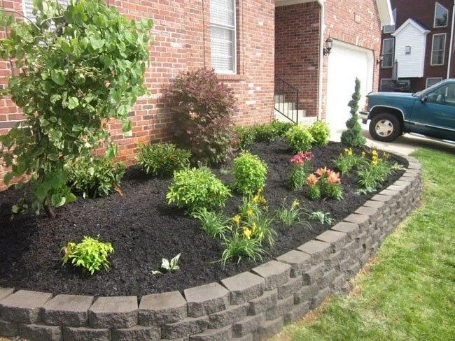 A well-landscaped garden bed in front of a brick house, enclosed by a dark gray retaining wall, filled with green plants, flowers, and dark mulch.