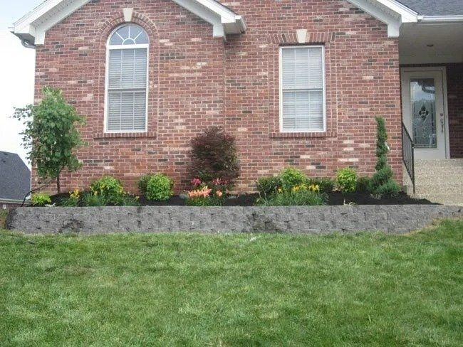 Brick house with a raised garden bed filled with flowers and shrubs, separated by a retaining wall from the green lawn.
