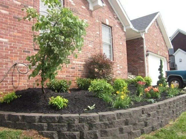 A brick house with a tiered garden bed. Dark mulch surrounds green plants, flowers, and a small tree. A garage is visible.