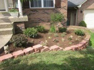 A brick-edged flower bed with mulch and green plants sits in front of a brick house with a garage.