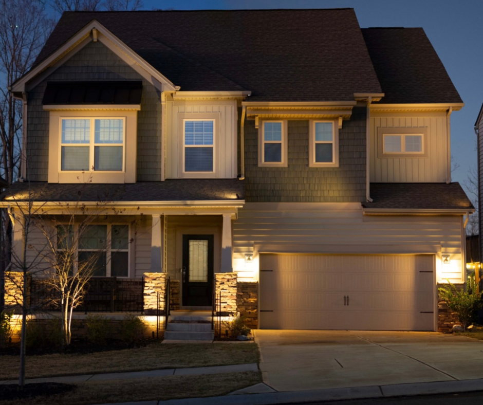A two-story house at dusk, lit by warm exterior lights. The facade is gray and tan with a black door.