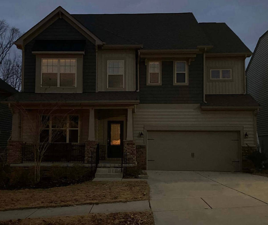 Two-story house with a garage, front porch, and lights on inside. Dark siding and beige details.