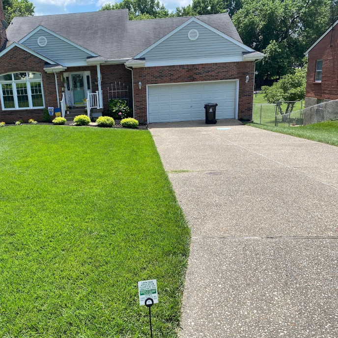 A well-manicured green lawn next to a concrete driveway leads to a brick house with a white garage door.