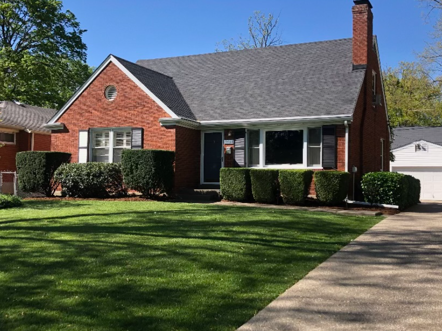 Brick house with a dark gray roof, green lawn, and trimmed bushes in front. A driveway is to the right.