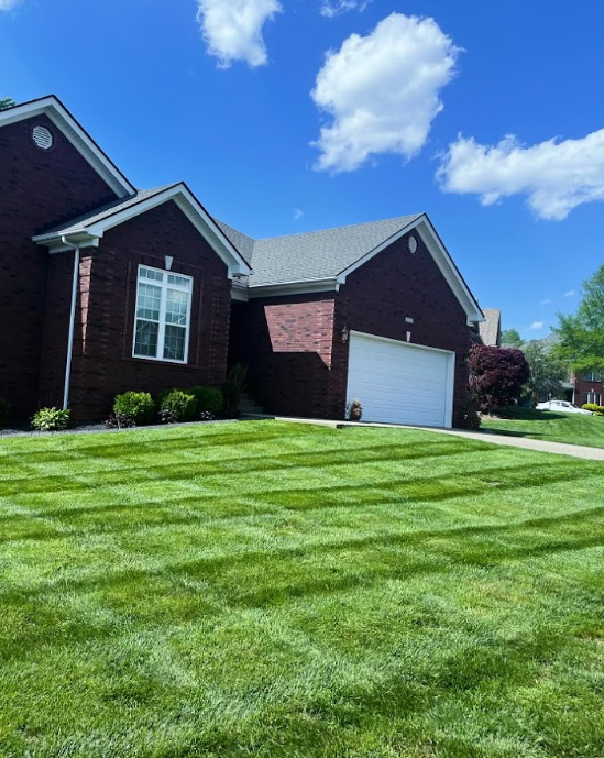 A brick house with a well-manicured lawn and striped grass under a blue sky with white clouds.