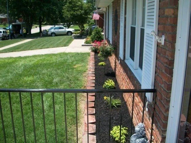 A house exterior with a brick facade, flower bed, and manicured lawn; a black metal fence is in the foreground.