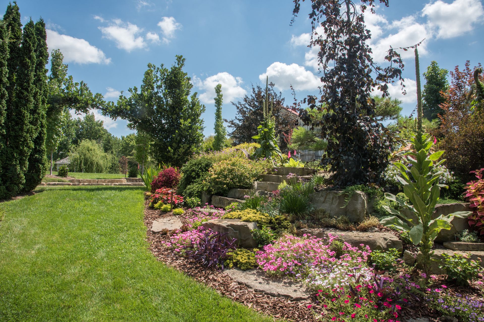 Lush garden with vibrant flowers, stone steps, and various trees under a partly cloudy blue sky.