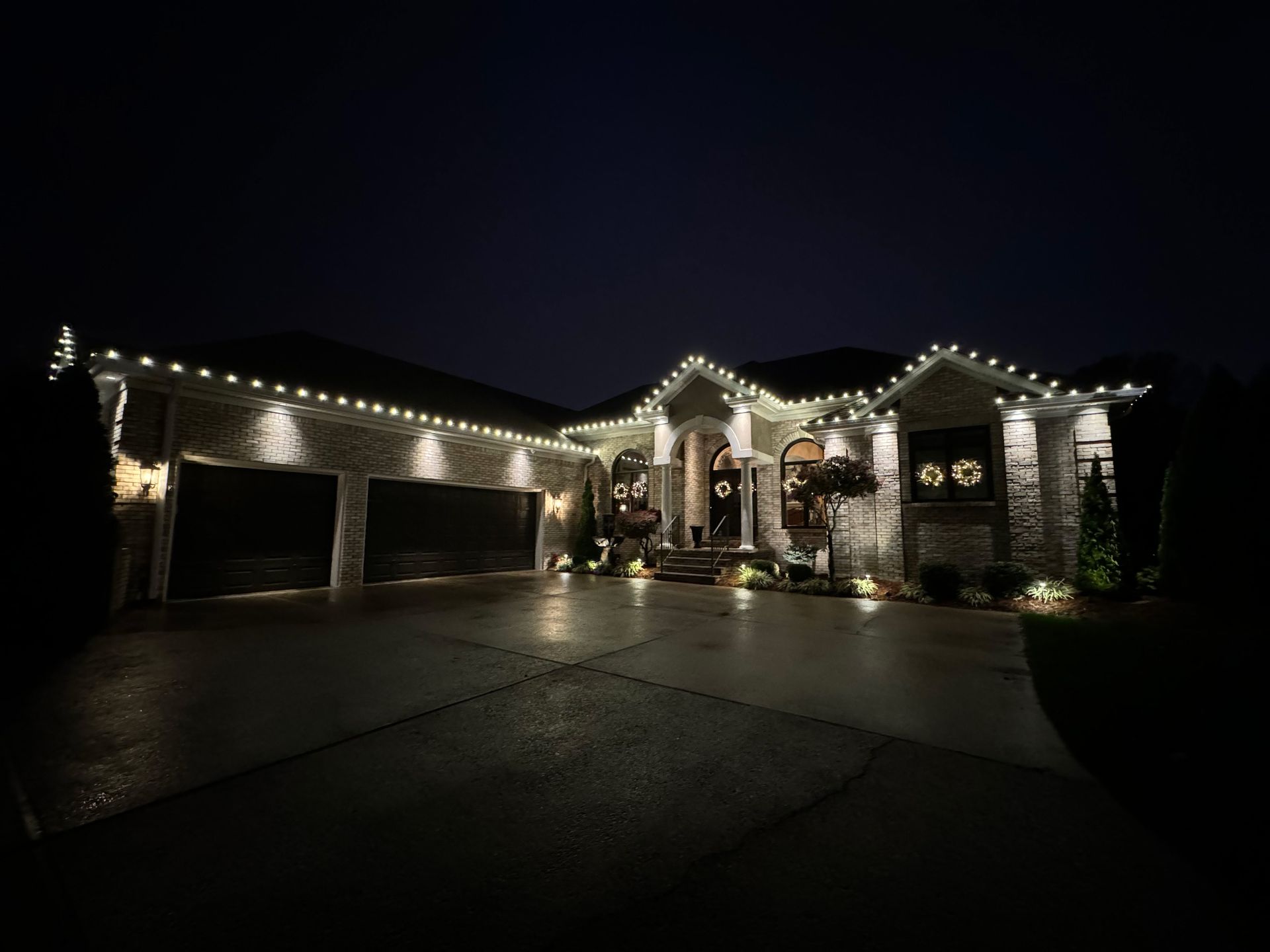 Nighttime view of a large house with Christmas lights along the roof and a lit driveway.