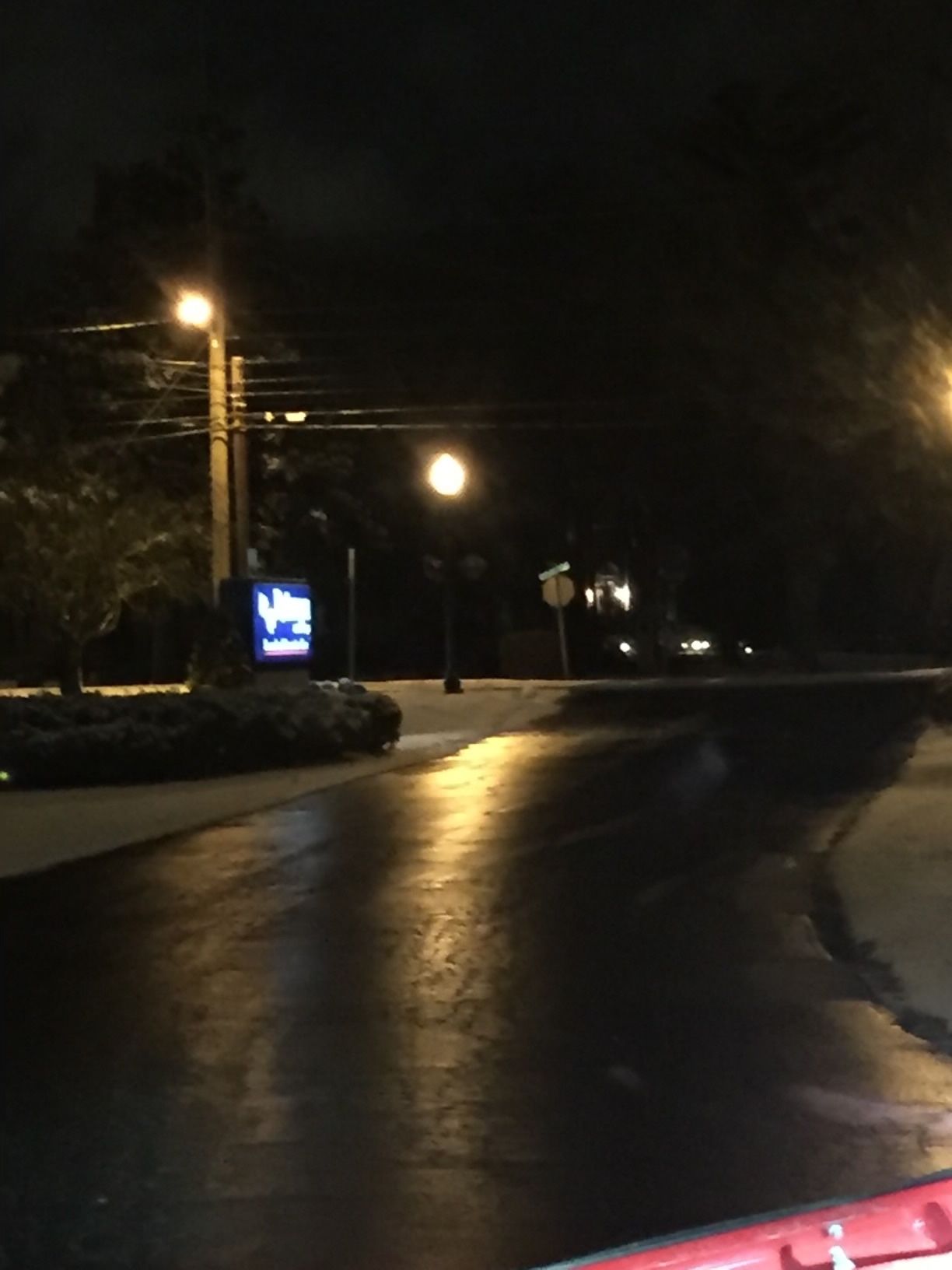 Dark, wet road at night with streetlights and a glowing sign along a quiet roadside.