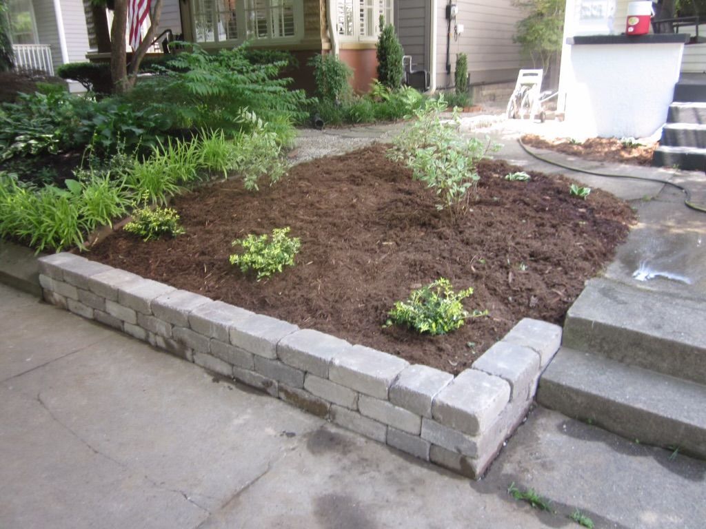 Stone retaining wall with a newly mulched garden bed in front of a house. Green plants are visible in the garden bed.