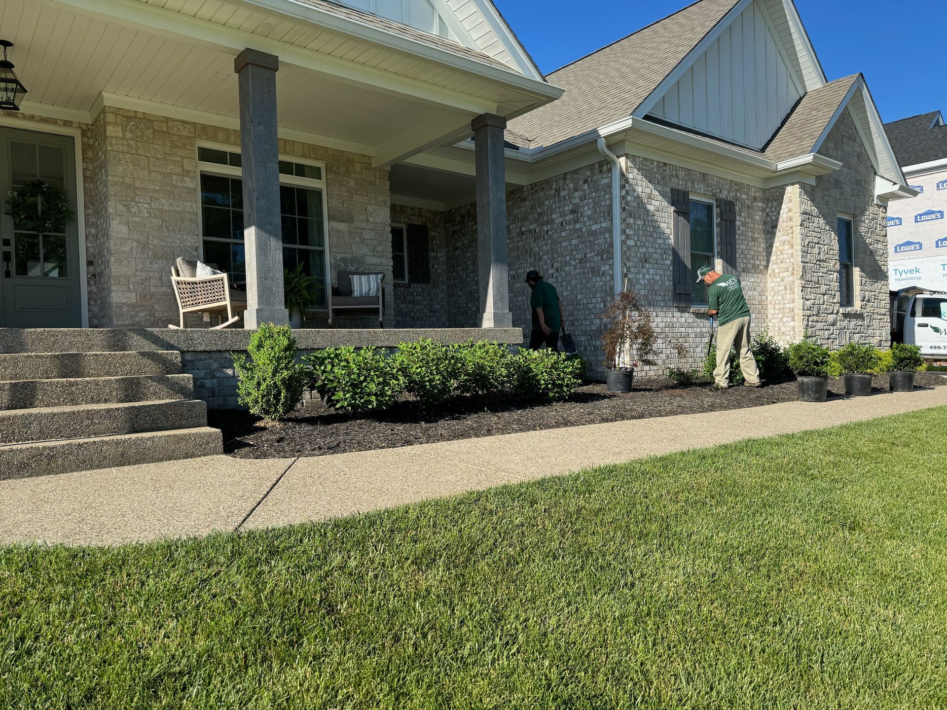 Landscapers planting bushes along the front of a house with a covered porch and steps. Green lawn and blue sky.